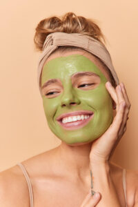headshot-pretty-pleased-young-woman-keeps-hands-face-looks-down-smiles-toothily-applies-green-nourishing-mask-has-combed-hair-wears-headband-poses-bare-shoulders-against-brown-studio-wall